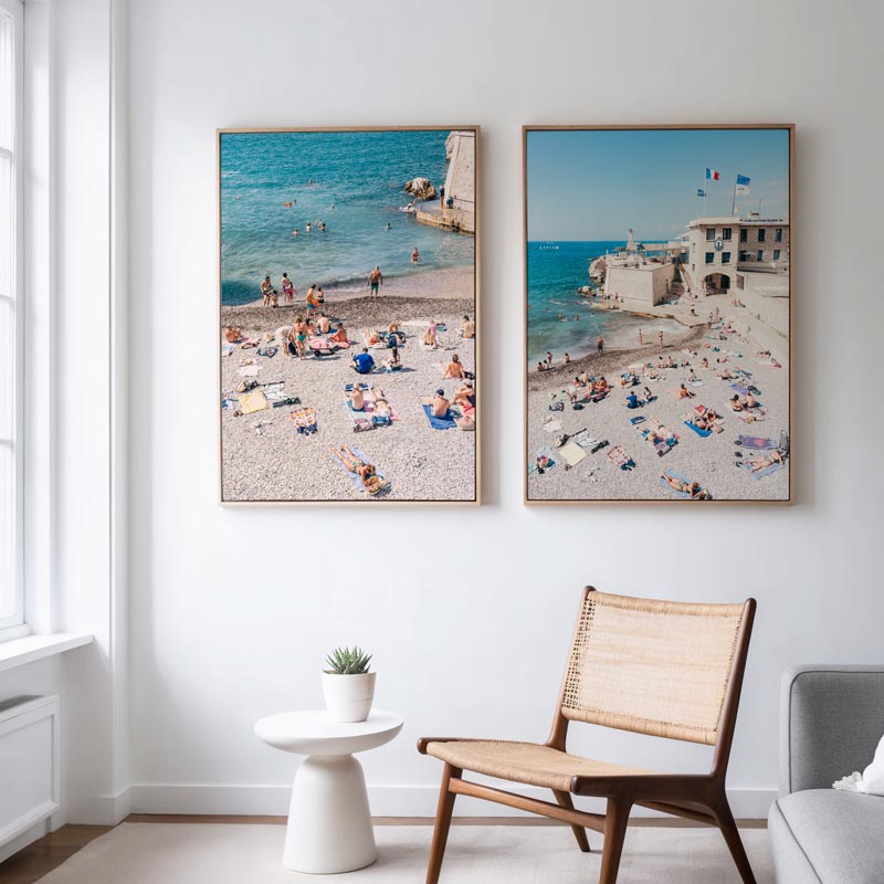 Two framed beach scene prints on a wall above a chair and small table.