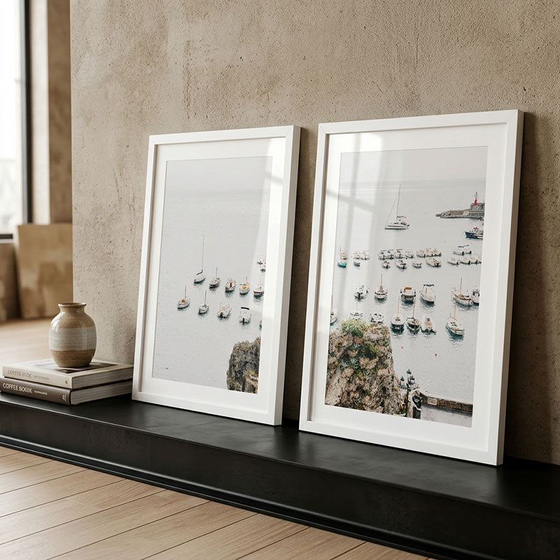 Two framed photographs of harbor scenes on a shelf against a beige wall.