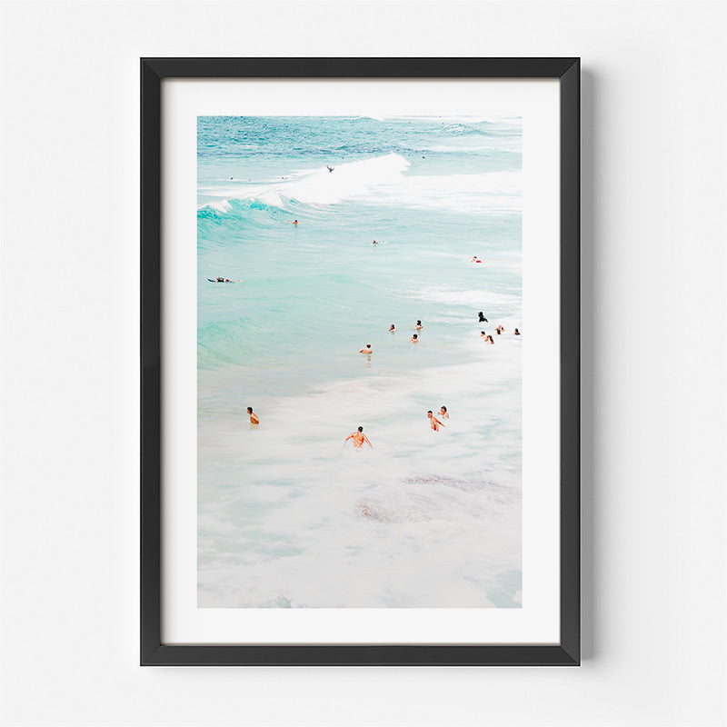 Framed artwork of a beach scene with people in the water on a white wall.