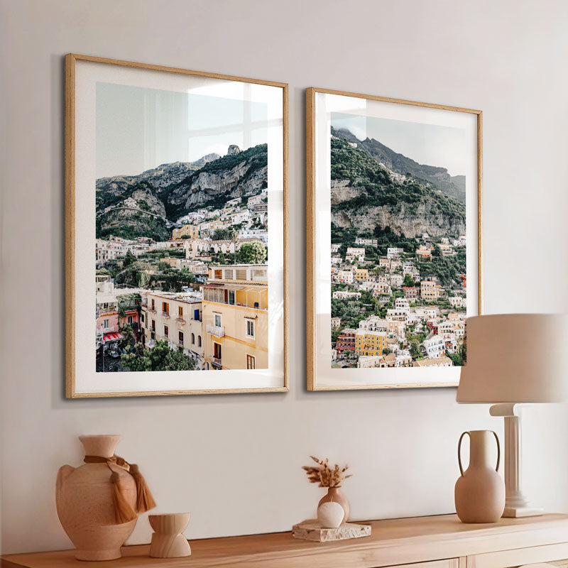 Two framed landscape prints of a mountainous village on a wall above a wooden console table.