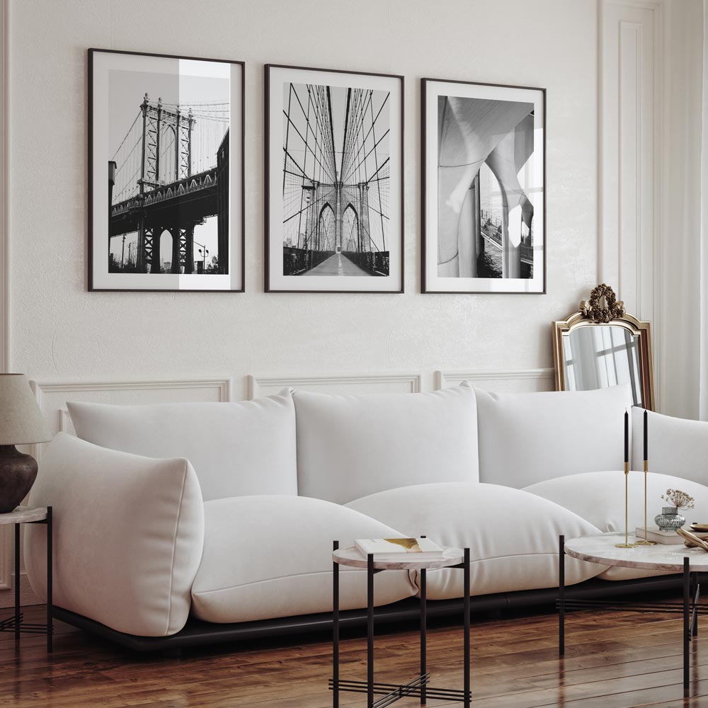 A set of three black and white photographs framed on a wall above a white sofa, with a view of the Brooklyn Bridge in the background.