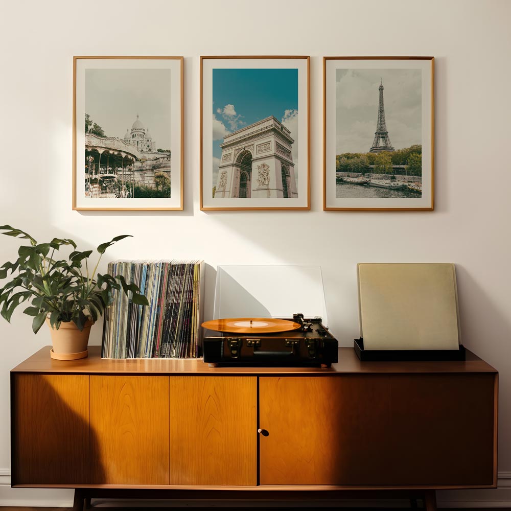 A set of three framed photographs hanging on a wall above a record player and a plant pot. The photographs feature iconic landmarks and a record player is placed in the foreground.