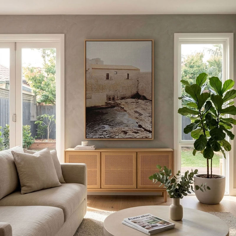 A large framed art print depicting a rustic stone building and pool, styled in a modern living room above a light wood credenza.