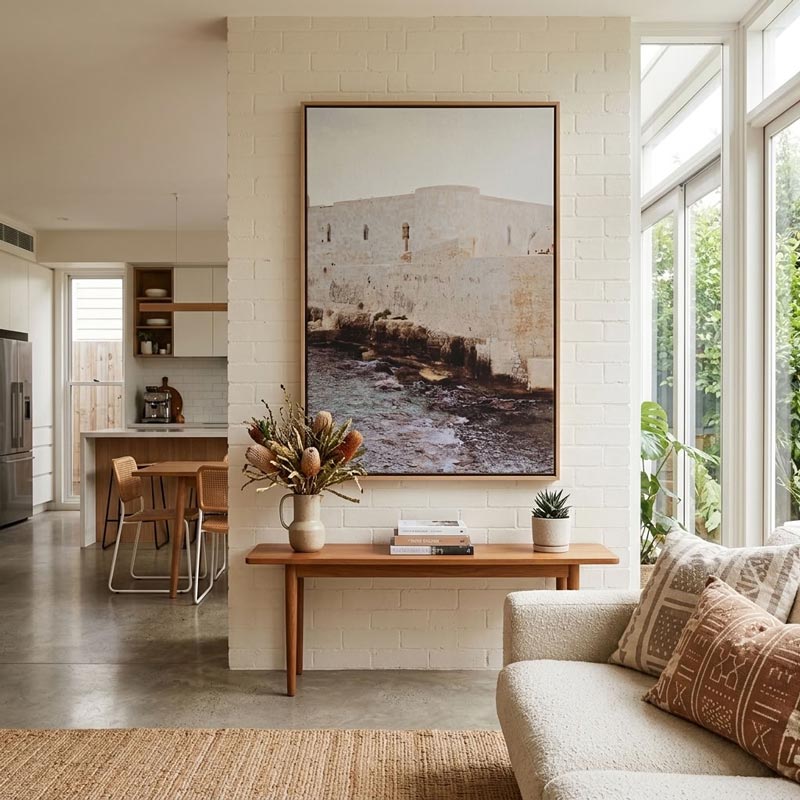 A large framed art print depicting a coastal stone wall and water, displayed on a white brick wall above a wooden console table in a modern, sunlit living space.