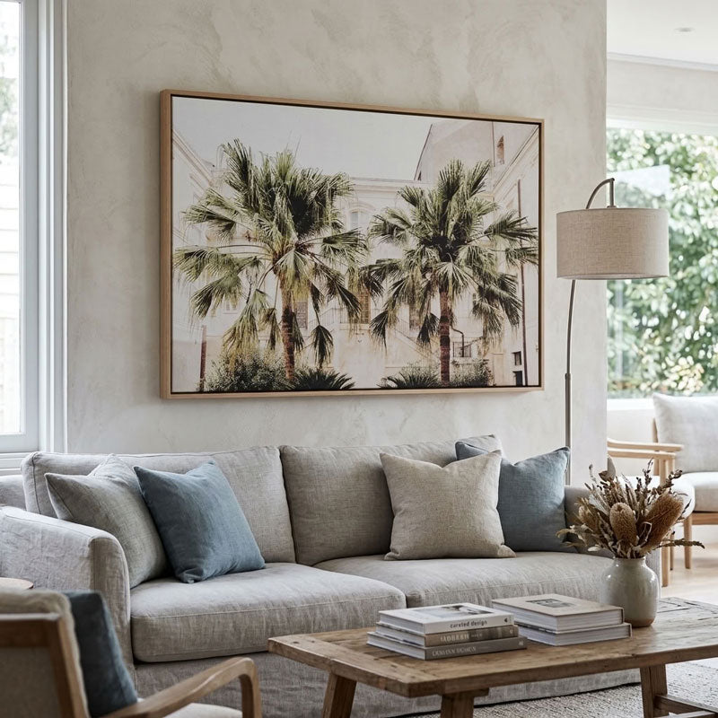 A large framed wall art print featuring two palm trees against a light building facade, displayed above a beige sofa in a bright, modern living room.