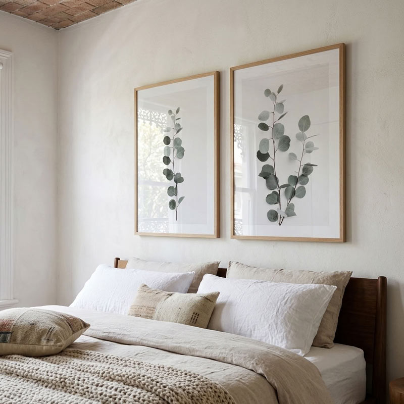 Two framed botanical prints with eucalyptus stems hang on a textured off-white wall above a bed with neutral-toned bedding and pillows.