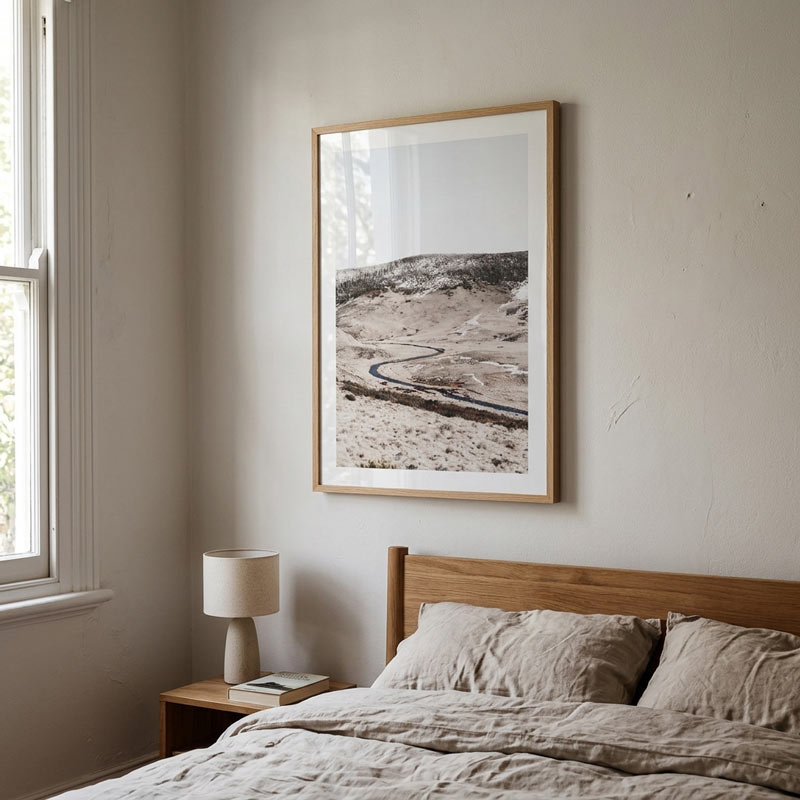 A framed landscape art print hangs on a beige wall above a wooden bed frame with neutral-colored bedding. The print depicts a winding road through a dry, sandy desert under a pale sky, enclosed in a light wood frame with a white mat.