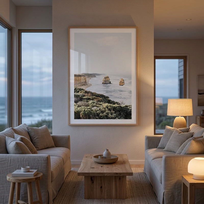 A large framed vertical art print of the Twelve Apostles sea stacks in Australia, displayed on a neutral beige wall in a modern, well-lit living room with gray sofas and a wooden coffee table.