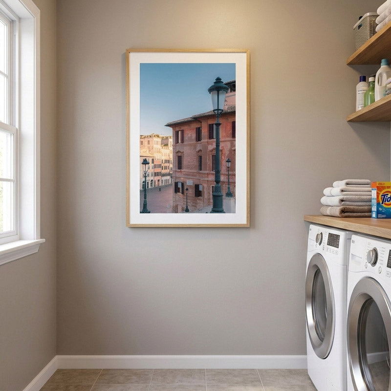 A framed vertical art print depicting a scenic street view of an old brick building and lampposts, displayed on a neutral gray wall in a laundry room setting next to a washing machine and dryer.