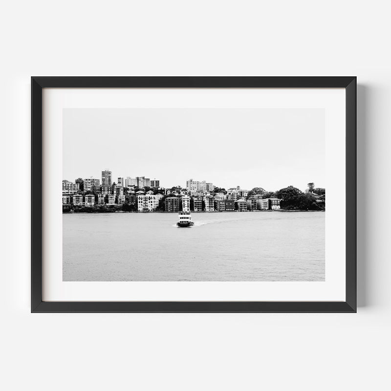 A framed black and white photograph of a waterfront city skyline with a small boat on the water, presented in a black frame with a white mat against a light gray background.