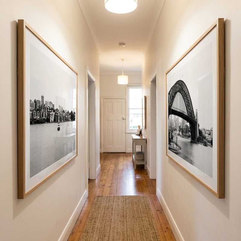 A long hallway with hardwood floors and cream walls, featuring two large framed black-and-white architectural photographs mounted on the walls. The left frame shows a city skyline over water, and the right frame shows a suspension bridge over water.