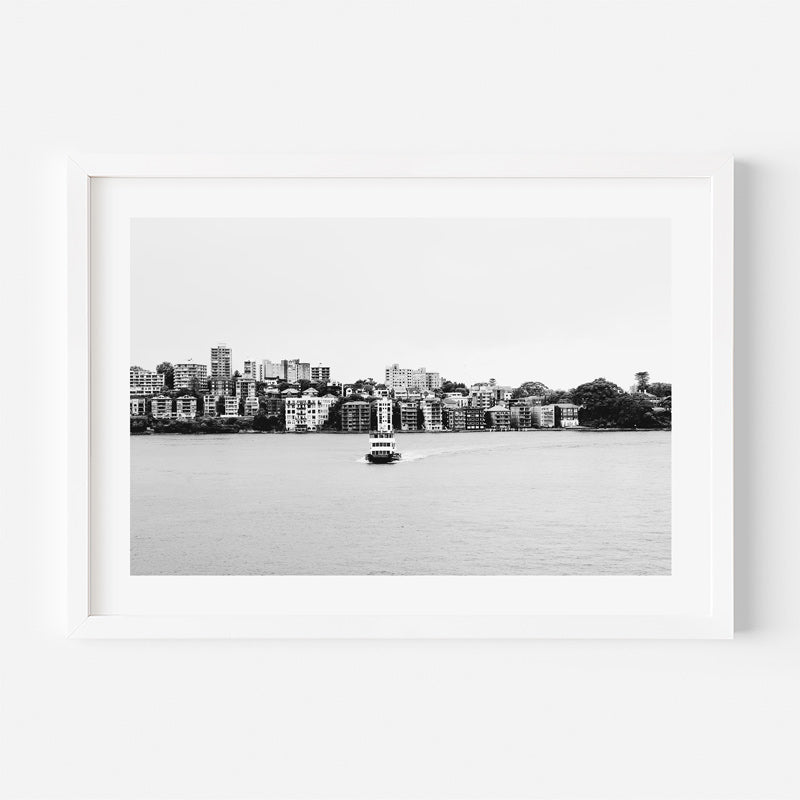 A framed black and white photograph of a waterfront city skyline with a small boat on the water, presented in a white frame against a light gray background.