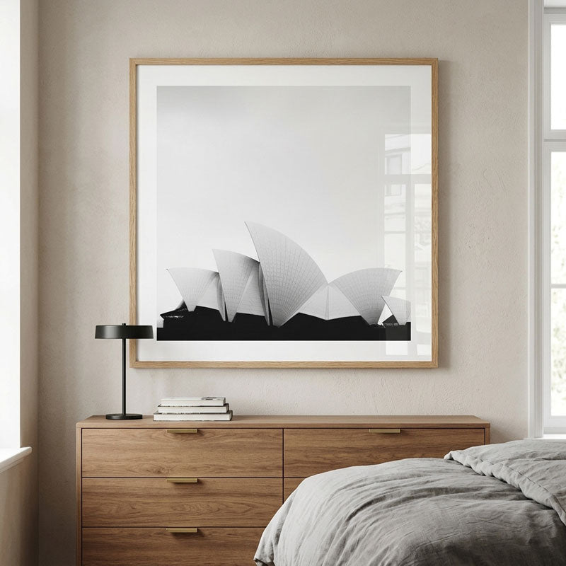 A large framed black and white art print of the Sydney Opera House, styled in a light wood frame, hanging on a beige wall above a wooden dresser in a bedroom setting.