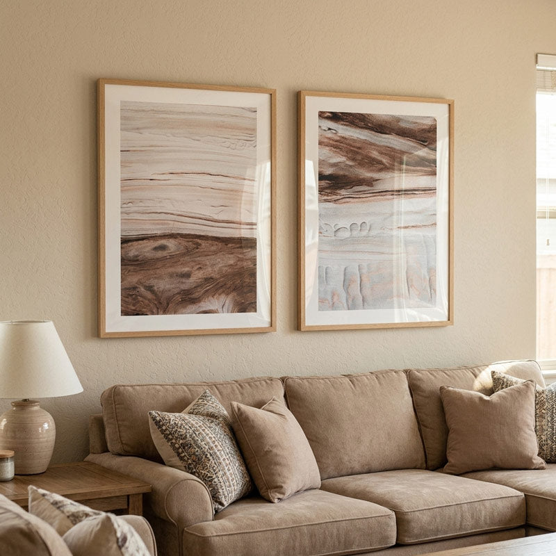 Two framed abstract art prints with light wood frames hang on a beige wall above a taupe sofa with matching pillows in a styled living room setting.