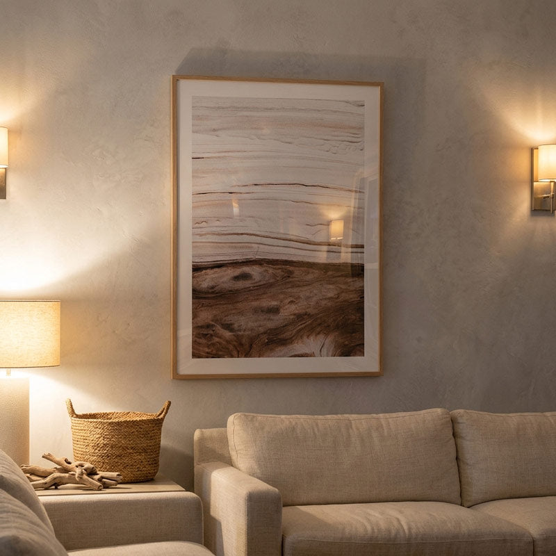 A framed abstract landscape art print with beige, brown, and white tones, hanging on a textured gray wall above a beige sofa in a warmly lit living room.