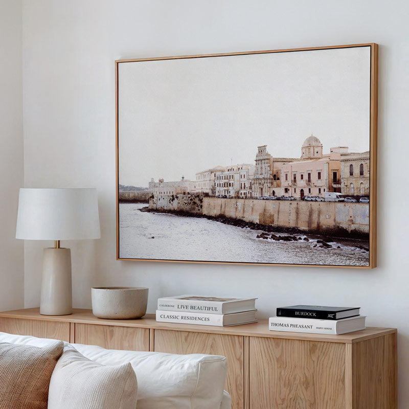 Living room with a large framed picture of a coastal scene, lamp, and books on a wooden cabinet.