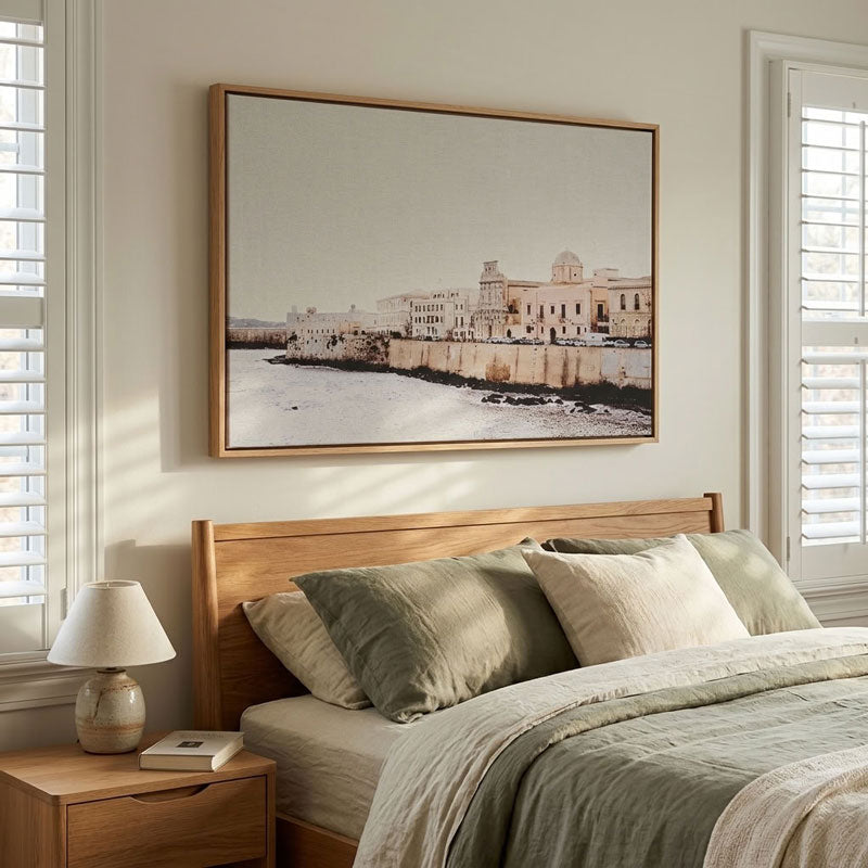A large framed art print depicting a coastal cityscape with historic buildings and a stone wall, hung on a beige wall above a wooden bed in a softly lit bedroom.