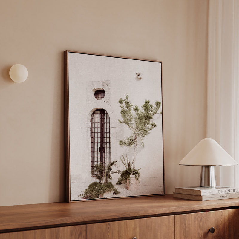 Framed artwork on a wooden shelf with a lamp and books in a room.