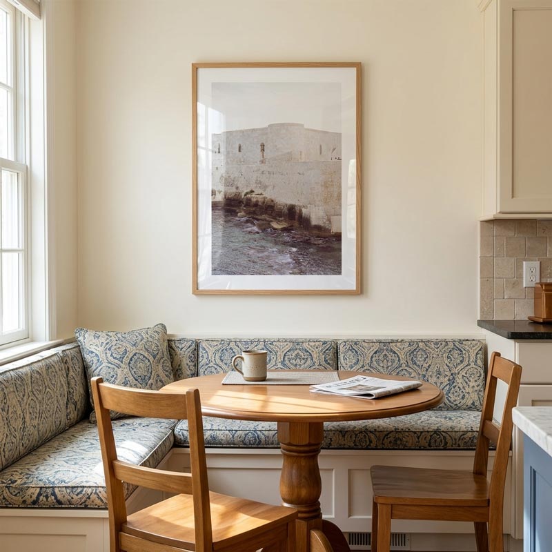 A framed art print depicting a stone building and water, hung on a cream wall above a wooden round dining table and a blue patterned banquette seating area in a bright kitchen.