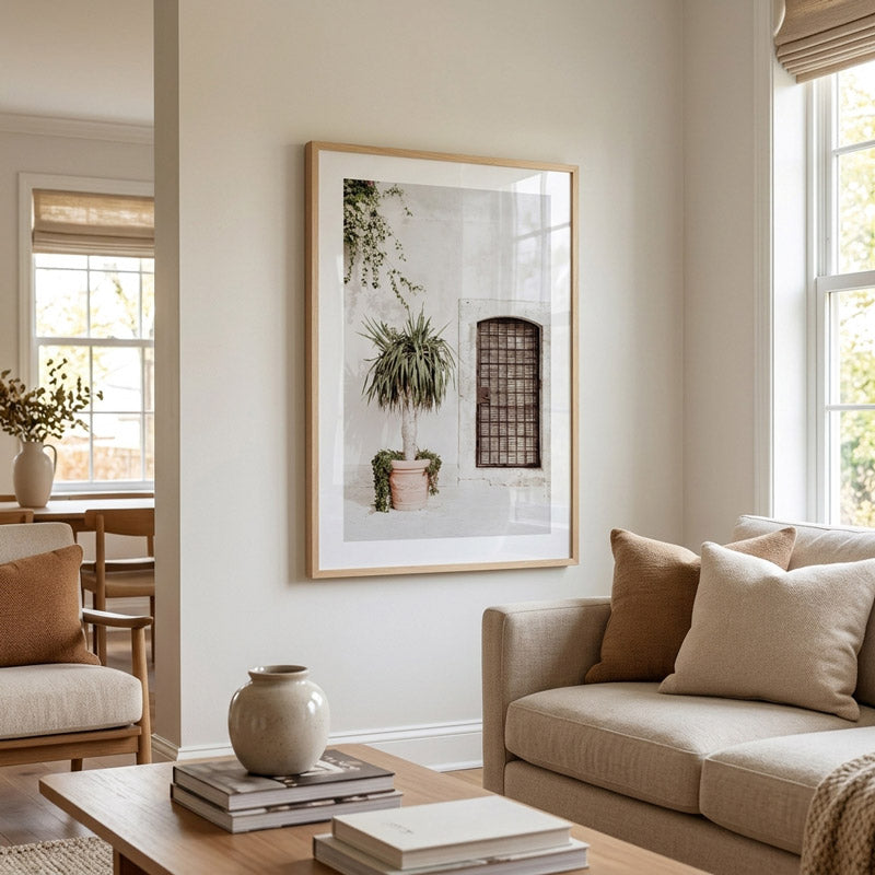 A framed art print featuring a minimalist architectural scene with a potted plant and window, displayed on a warm beige wall in a modern living room setting.