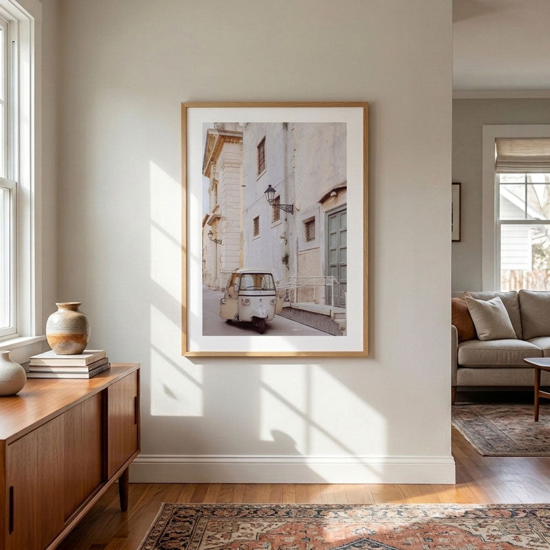 A framed art print depicting a vintage white car parked on a narrow street between light-colored buildings, hung on a neutral wall in a modern living room setting.