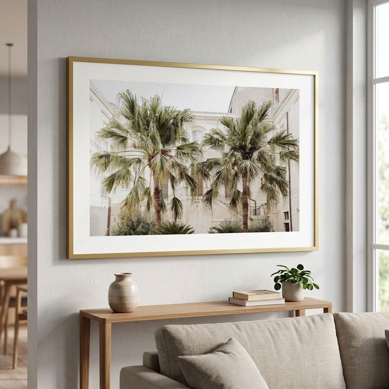 A framed art print featuring two palm trees in front of a white building, styled on a light gray wall above a wooden console table with a sofa in a modern living room setting.