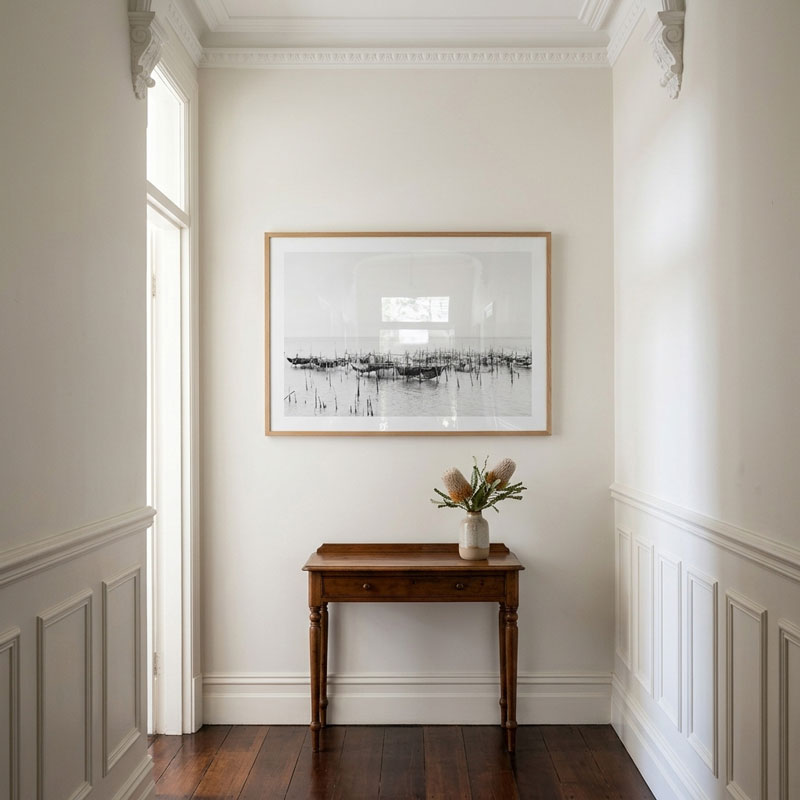 A framed black and white landscape art print hangs on a white wall in a bright hallway above a wooden console table with a vase of flowers.