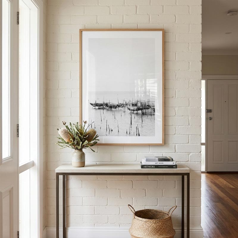 A framed black and white art print of boats on water, in a light wood frame, displayed on a white brick wall above a console table in a bright, modern room.