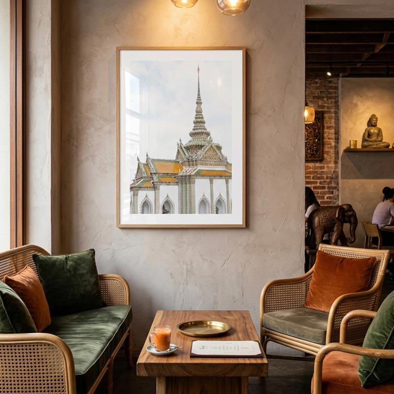 A framed art print depicting a Thai temple tower hangs on a textured beige wall in a cozy, modern living room setting with woven chairs and a wooden coffee table.