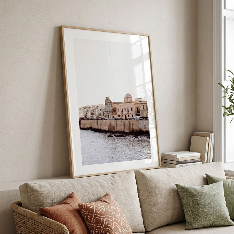 A large framed art print of a coastal cityscape with historic buildings and water, leaning against a beige wall above a light-colored sofa with decorative pillows.