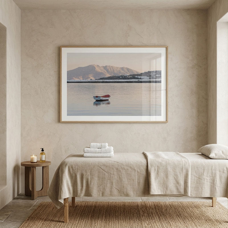 A framed landscape art print of a small boat on a calm lake with mountains in the background, displayed on a textured beige wall above a spa-style treatment table in a serene room.