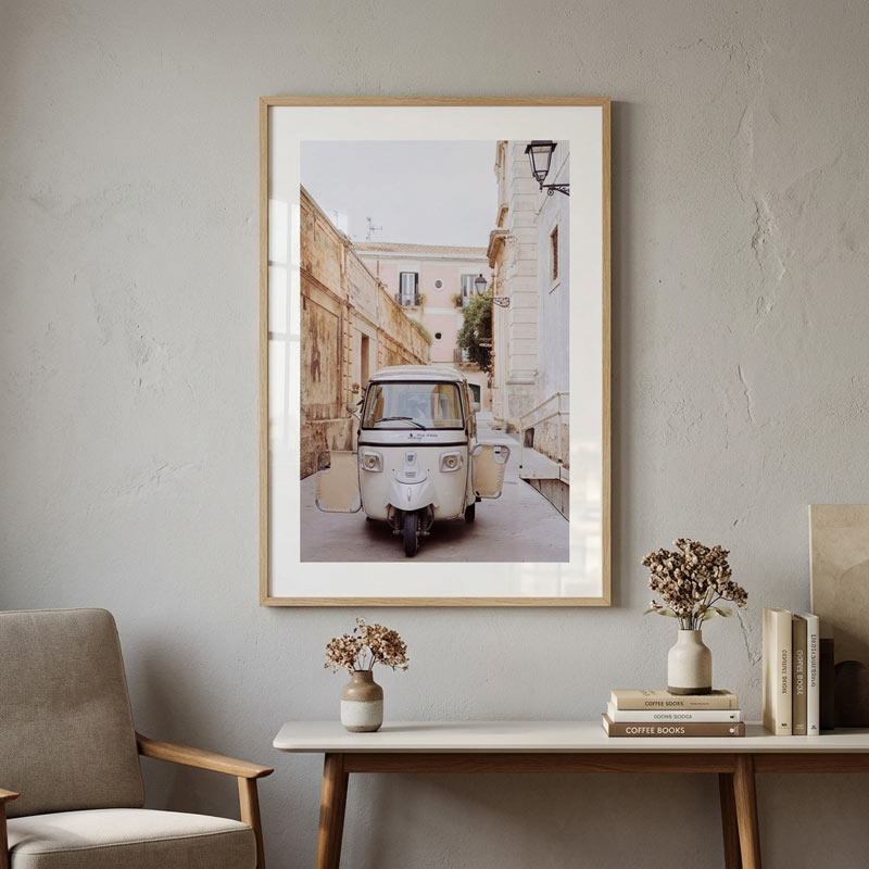 A framed art print of a vintage white scooter parked in a narrow European street, displayed on a textured light gray wall above a console table with books and vases.