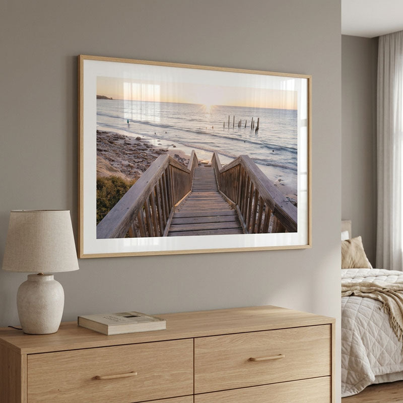 A framed wall art print showing a wooden boardwalk leading to the ocean at sunset, displayed above a light wood dresser in a modern bedroom setting.