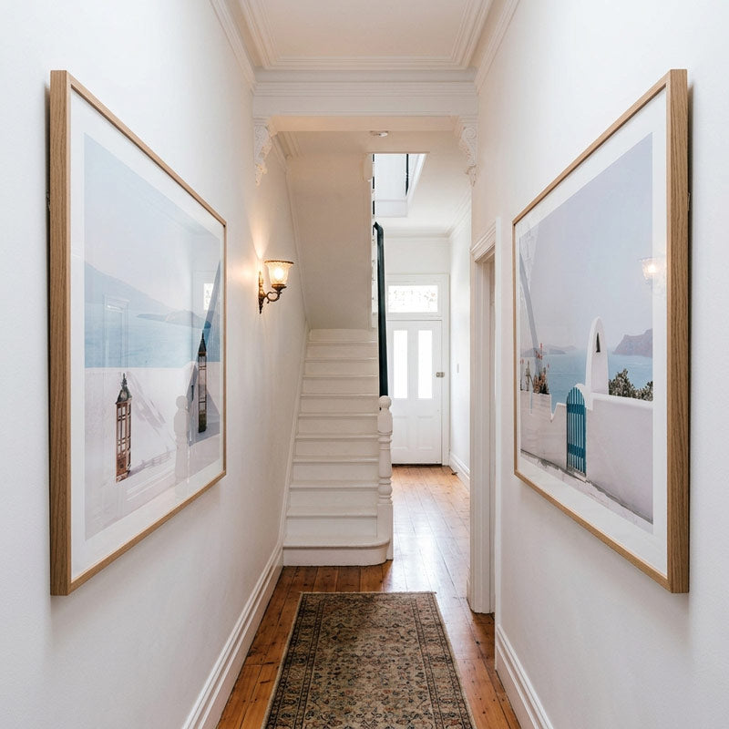 A long hallway with white walls, wood flooring, a staircase in the distance, and two large framed art prints hanging on either side. The prints depict coastal scenes with white buildings, blue doors, and soft blue tones.