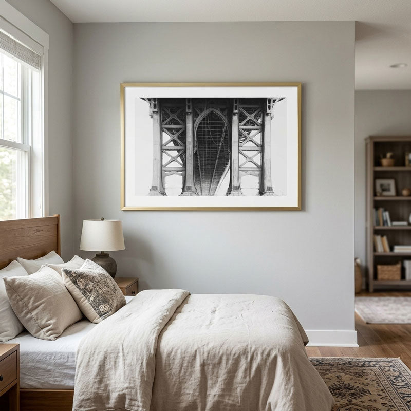 A framed black and white art print of the Manhattan Bridge hanging on a light gray wall above a neatly made bed in a modern, neutral-toned bedroom.