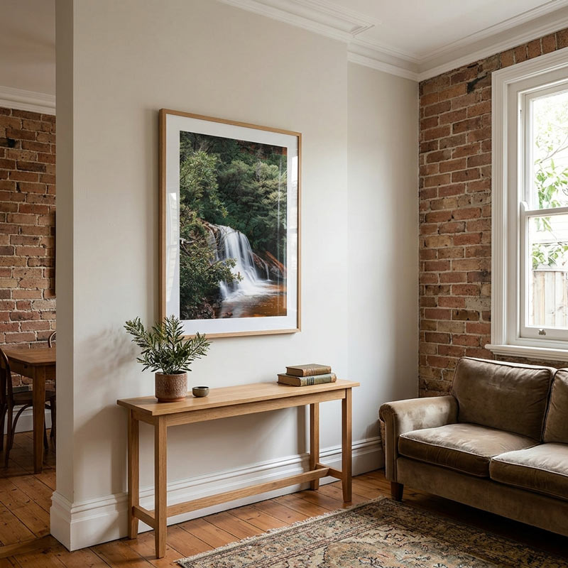 A framed vertical landscape art print of a waterfall in a forest, displayed on a white wall in a styled living room with a wooden console table, brick walls, and a sofa.