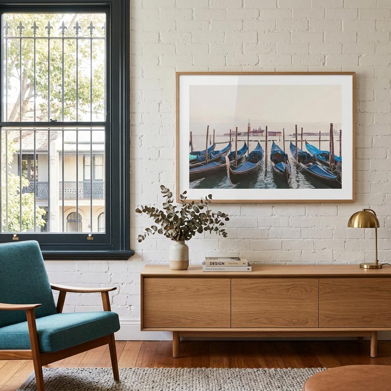 A framed art print depicting gondolas in Venice, displayed on a white brick wall above a wooden console table in a modern living room setting.
