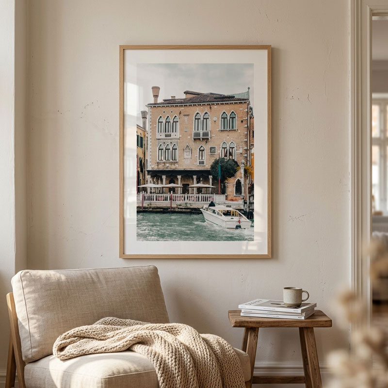 A framed art print depicting a Venetian canal scene with historic buildings and a boat, displayed on a beige wall above a light-colored armchair with a knitted throw blanket and a small wooden side table with books and a cup.