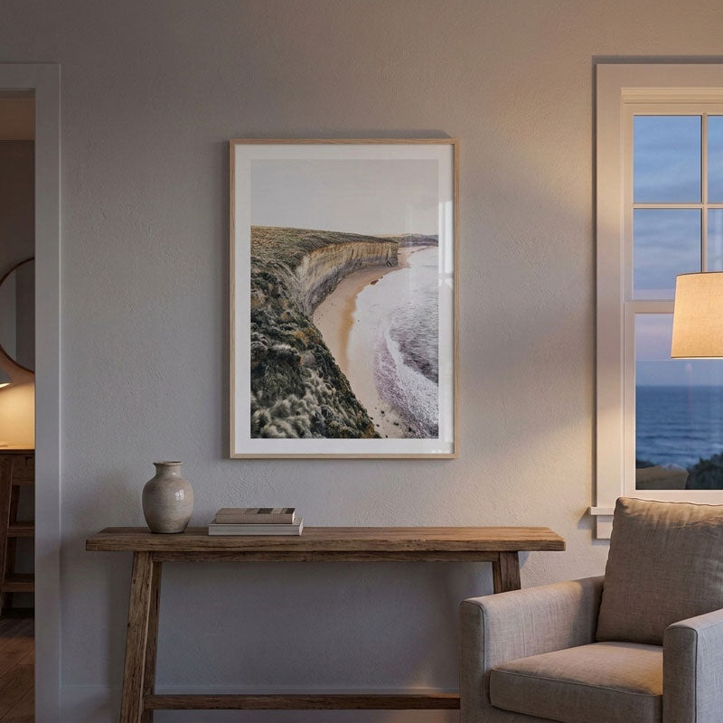 A framed coastal landscape art print hangs on a gray wall above a rustic wooden console table in a styled living room setting.