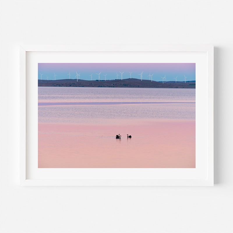 Framed artwork of a landscape with wind turbines and a pink sunset over water.