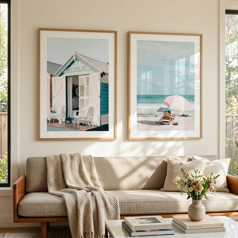 Two framed beach-themed art prints hang on a beige wall above a light beige sofa in a styled living room. The left print shows a small white beach hut with blue shutters, and the right print shows a beach scene with a pink umbrella and lounge chair.