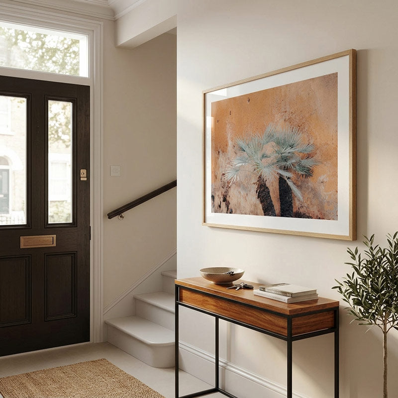 A framed abstract art print featuring a blue-green succulent on a textured beige and orange background, displayed on a white wall in a modern entryway above a wooden console table.