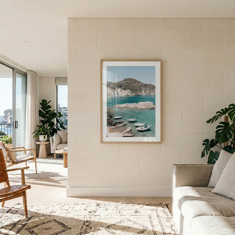 A framed art print depicting a scenic coastal bay with turquoise water, boats, and rocky cliffs, displayed on a light beige brick wall in a modern, sunlit living room.