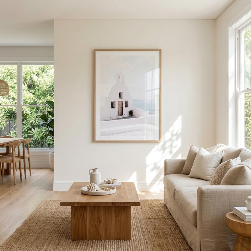 A framed art print depicting a white church with a dome and a small door, hung on a warm beige wall in a bright, modern living room with a wooden coffee table and a beige sofa.
