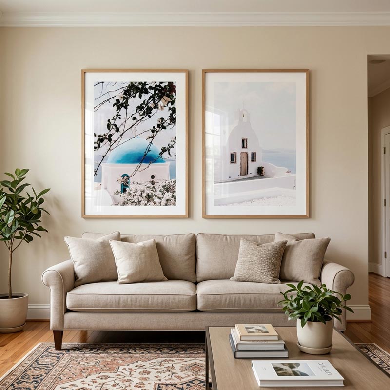 Two framed art prints depicting Santorini scenes hang on a beige wall above a light beige sofa in a styled living room. The left print features a white building with a bright blue dome under tree branches, and the right print shows a white church building with a pale blue sea and sky.
