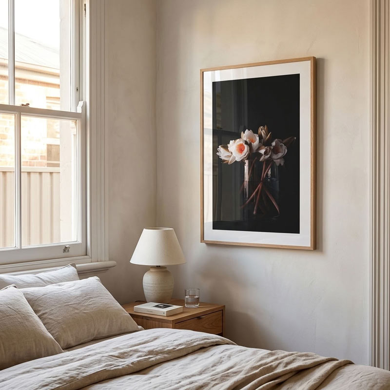 A framed floral art print hangs on a beige wall above a bed in a softly lit bedroom. The print shows white and pink flowers against a dark background, enclosed in a light wood frame with a white mat.