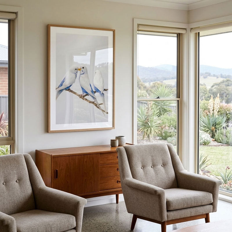 A framed art print featuring two blue and white birds perched on a branch, displayed on a light beige wall in a modern living room setting with two upholstered armchairs and a wooden sideboard.