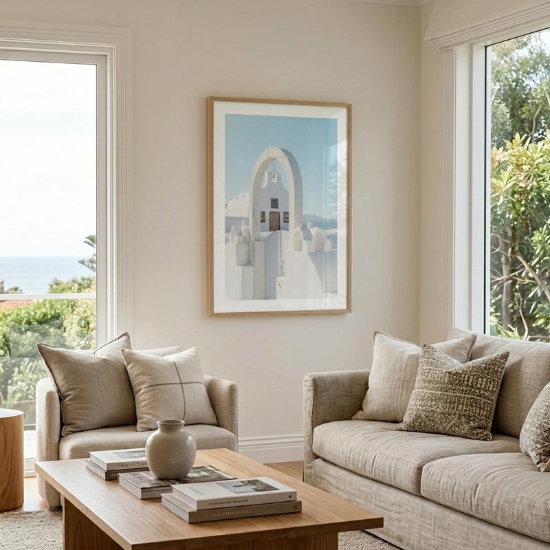 A framed art print featuring a Mediterranean-style white archway building under a light blue sky, displayed on a beige wall in a bright, modern living room with a sofa, armchair, and wooden coffee table.