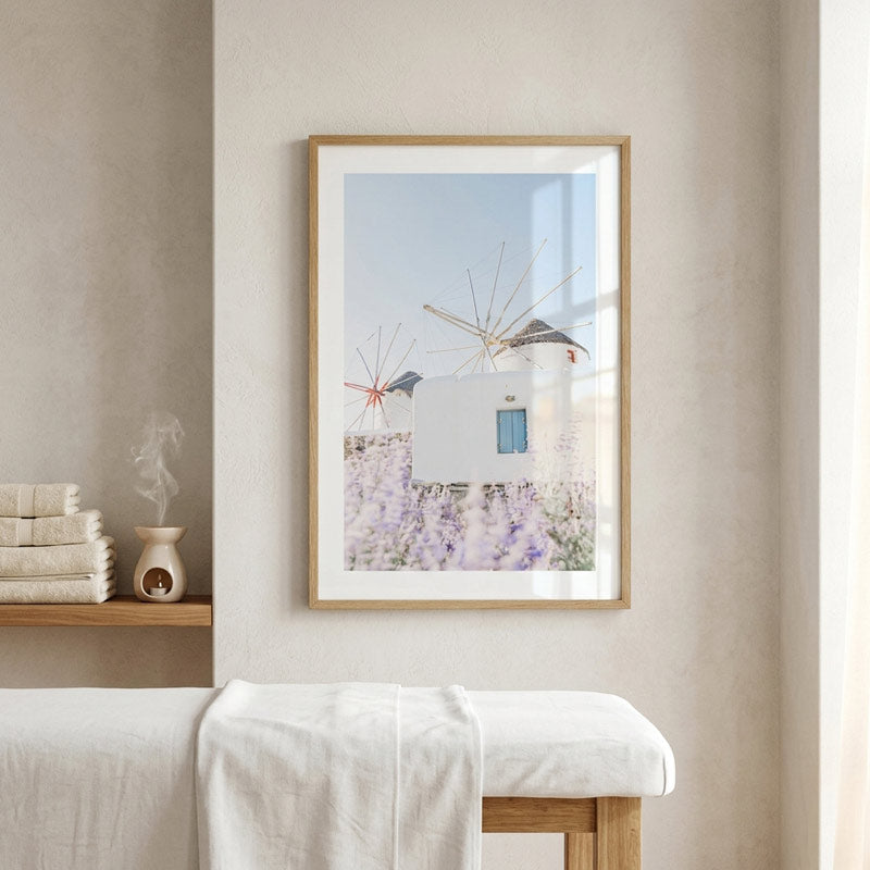A framed art print depicting a Mediterranean scene with white houses, lavender fields, and windmills, hanging on a beige wall above a white massage table in a spa-like setting.