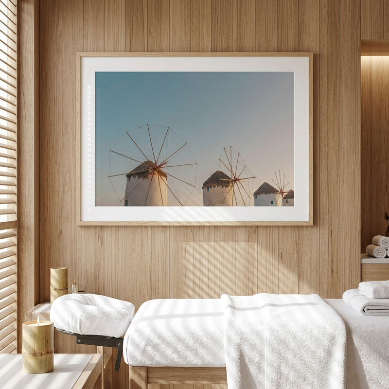 A framed art print depicting traditional wind mills under a clear blue sky, displayed on a light wood-paneled wall above a spa treatment table with white towels.